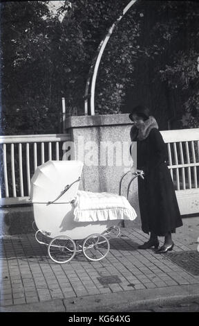 @ Années 1920, une jeune mère à la mode du jour portant un manteau de fourrure garni et chapeau avec son ressort élégant quatre roues pram dehors sur un trottoir avec une courtepointe couvrant. Banque D'Images