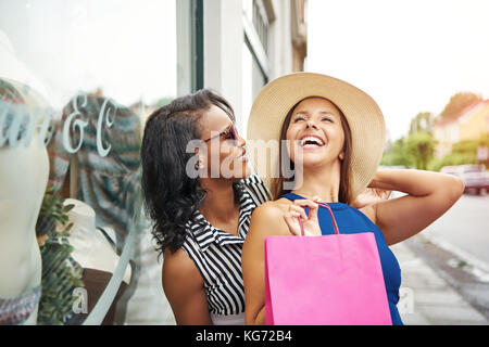 Paire d'amis multiraciale en robes sans manches, chapeau et des lunettes de window shopping together Banque D'Images