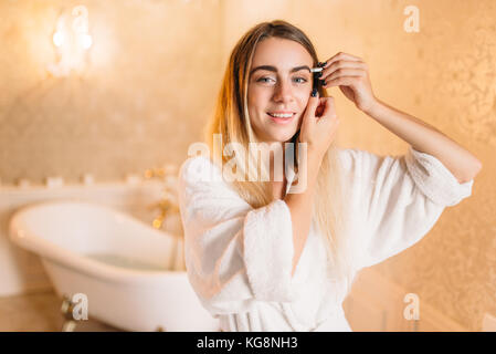 Happy woman in white peignoir faisant un miroir dans la salle de bains, soins visage dans la salle de bains. Soins du corps et de l'hygiène, les soins de santé Banque D'Images