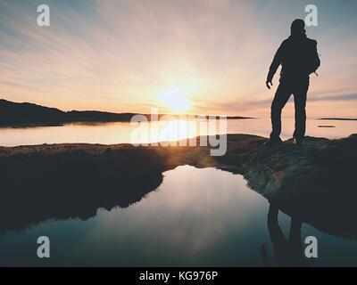 Grand randonneur dans dark sportswear avec sac à dos sportif se dresse sur falaise au-dessus de la mer. L'homme profitez incroyable coucher du soleil à l'horizon avec orange ciel rose avec des nuages. ma Banque D'Images