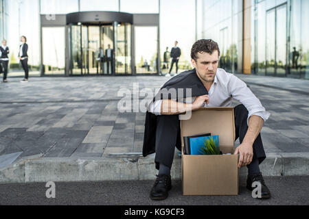 Tiré business man sitting frustré et contrarié dans la rue près de l'immeuble de bureaux de fort de ses affaires. Il a perdu travail Banque D'Images