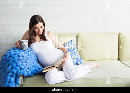 Jolie Jeune femme enceinte à l'intérieur assis sur un canapé et de la lecture Banque D'Images