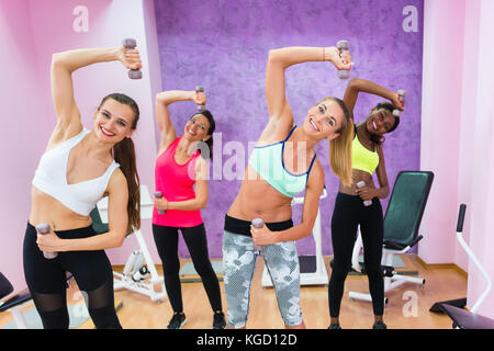 Quatre femmes gaies holding dumbbells tout en faisant des exercices pour les bras et les muscles abdominaux latéraux au cours de conditionnement physique de groupe, classe dans un club de santé moderne Banque D'Images