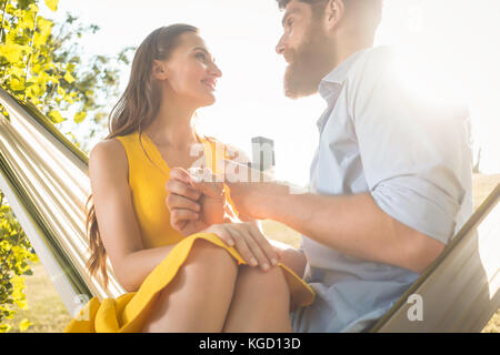 Bas côté angle de vue d'un jeune homme gai de faire une proposition de mariage à sa belle petite amie, tout en se balançant dans un hamac à l'extérieur dans un Banque D'Images