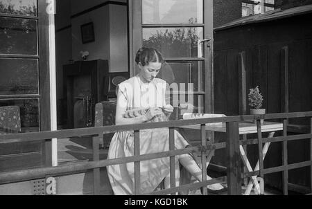 1940, la Grande-Bretagne, jeune femme une robe assis seul sur une véranda accueil tricot. Le tricot à la main était un passe-temps populaire pour les femmes pendant cette période de guerre. Banque D'Images