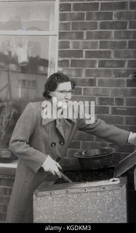 Années 1950, historique, dame en manteau à l'aide d'une pelle pour remplir un petit bassin avec du charbon de la hauteur de la taille de l'unité de stockage en acier à l'extérieur, England, UK. Banque D'Images