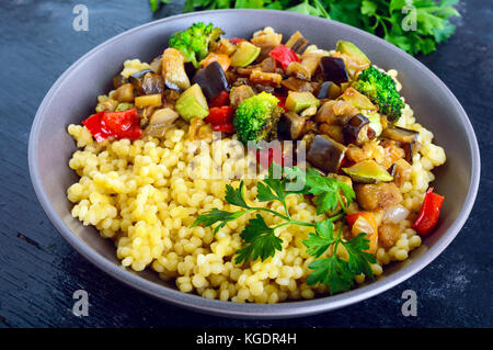 Un régime alimentaire léger plat végétalien : couscous et légumes (courgettes, aubergines, carottes, poivrons, oignons, persil) sur un fond noir. Banque D'Images