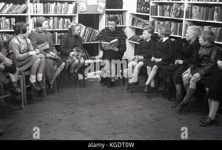 Années 1950, historiques, l'heure du conte à l'école....photo montre une enseignante en train de lire à partir d'un répertoire pour un groupe d'enfants de l'école britannique tranquillement installé sur des chaises dans un coin de la bibliothèque. Banque D'Images