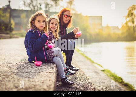 Family eating ice cream, assis sur le bord de la rivière Ljubljanica. mère et filles jumelles sont assis près d'une rivière, manger le dessert avec rivière et s Banque D'Images