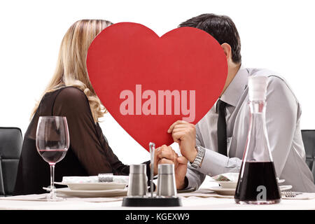 Jeune couple assis à une table de dîner et de s'embrasser derrière un cœur rouge isolé sur fond blanc Banque D'Images