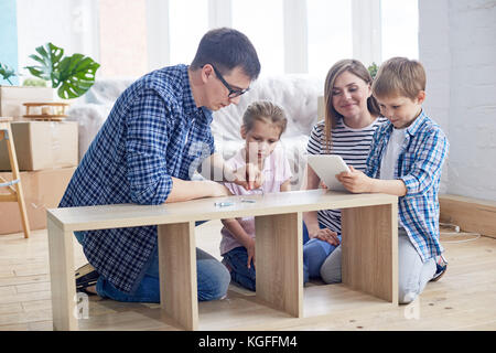 Famille de quatre jeunes amoureux réunis au salon de l'appartement neuf et enveloppé dans l'assemblage armoire, pile de cartons de déménagement sur backgroun Banque D'Images