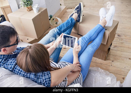 Vue arrière du jeune couple charmant à regarder leurs émissions préférées sur tablette numérique série et assis sur un canapé confortable tout en pause de trucs dans l'emballage Banque D'Images