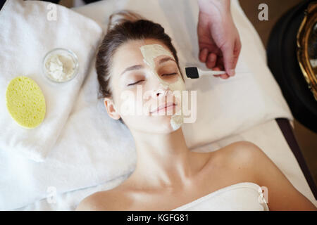 Vue de dessus portrait de belle jeune femme dans SPA, couché sur la table de massage avec les yeux fermés tout en appliquant cosmetologist au visage Masque visage Banque D'Images