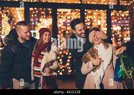 Les jeunes professionnels quatre amis dans shopping passe devant la fenêtre du centre commercial, et l'homme conserve sa petite amie alors qu'elle les yeux couverts de donner un cadeau. Banque D'Images