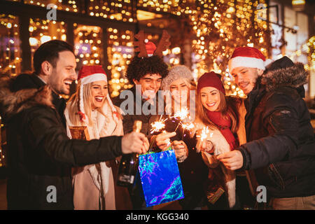 Cheerful six jeunes amis s'amusant dans le centre commercial de la ville dans la nuit de Noël, alors que l'aide de baguettes, avec vitrine lumineuse en arrière-plan. Banque D'Images