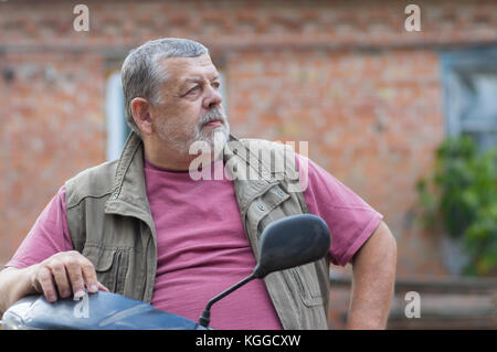 Beau portrait de senior homme barbu assis sur un scooter contre brick wall Banque D'Images