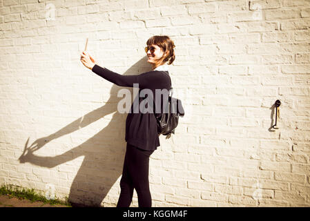 Jeune femme vêtue de noir avec un petit sac à dos porter des lunettes de soleil de sourire et de prendre un en selfies avant d'un mur en brique blanche en arrière-plan Banque D'Images