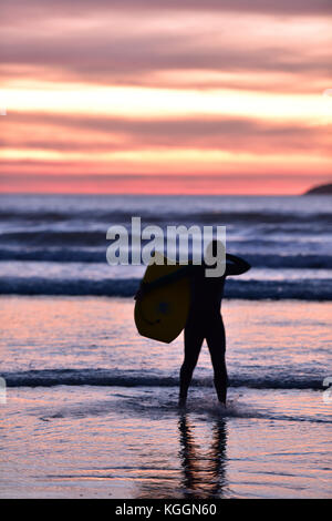 Un surfeur sur la plage au coucher du soleil en silhouette à polzeath en Cornouailles du nord transportant une planche et pagaie dans les vagues et surfer sur les disjoncteurs. Banque D'Images