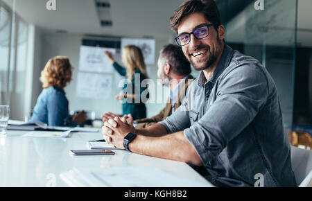 Smiling designer assis en salle du conseil d'administration lors de la présentation. Jeune homme assis au bureau et à la recherche à l'appareil photo. Banque D'Images