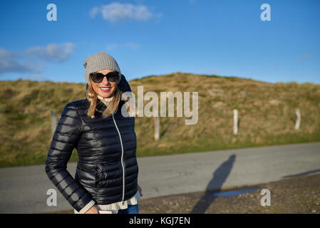 Femme à la mode de porter une veste en cuir, bonnet de laine et lunettes debout sur une route rurale dans la lumière du soir avec copie espace smiling at the camera Banque D'Images