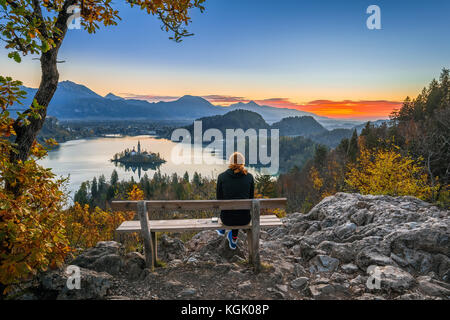 Bled, Slovénie - une femme coureur de cheveux rouge se détendant et profitant de la belle vue d'automne et du lever de soleil coloré du lac Bled assis sur une colline benc Banque D'Images