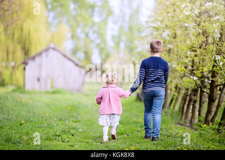 Jolie petite fille en robe rose et le garçon en bleu jeans pull et marcher ensemble loin d'un appareil photo, se tenant la main et à l'arrière Banque D'Images