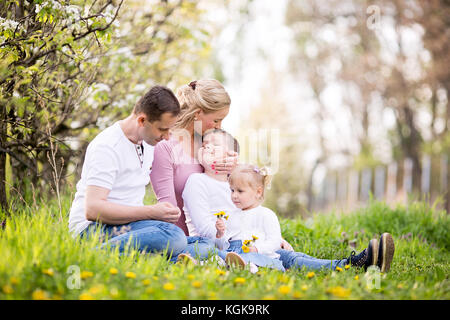 Quatre États heureux jeune famille prévues printemps après-midi ensemble à l'extérieur en verger, assis dans une herbe Banque D'Images