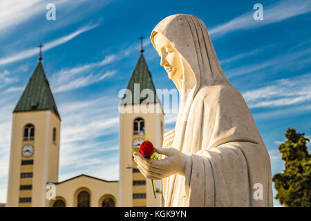 Statue de la Vierge Marie tenant une rose rouge alors que l'église Saint Jacques de Medjugorje est en arrière-plan Banque D'Images