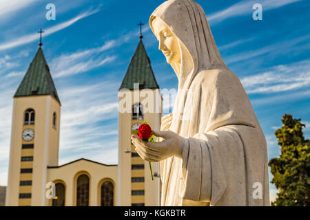 Statue de la Vierge Marie tenant une rose rouge alors que l'église Saint Jacques de Medjugorje est en arrière-plan Banque D'Images