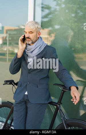 Businessman with bicycle talking on mobile phone debout contre l'édifice de verre Banque D'Images