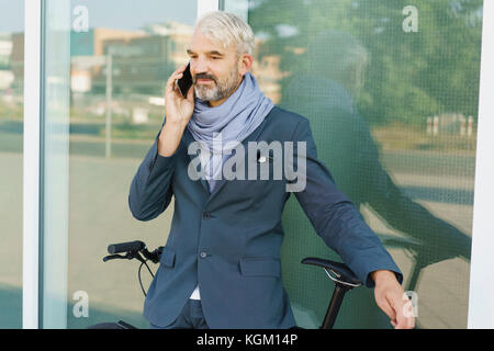 Confident businessman with bicycle talking on mobile phone debout contre l'édifice de verre Banque D'Images