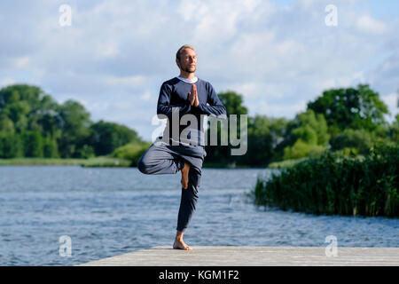 Homme mûr pratiquant le yoga posture de l'arbre sur la jetée par le lac against sky Banque D'Images