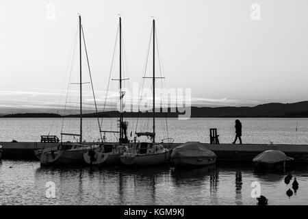 Un port sur un lac au coucher du soleil, avec des bateaux et une femme parlant au téléphone tout en marchant Banque D'Images