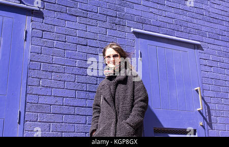 Femme avec des lunettes de hipster, debout devant un mur de brique violet lumineux. Le port de la veste en laine de couleur noir et l'écharpe. Banque D'Images