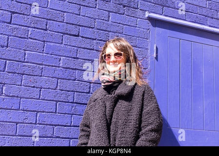 Femme avec des lunettes de hipster, debout devant un mur de brique violet lumineux. Le port de la veste en laine de couleur noir et l'écharpe. Banque D'Images