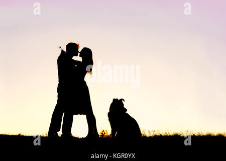 La silhouette d'un jeune couple marié dans l'amour, serrant au coucher du soleil à côté de leur chien de compagnie à l'extérieur sur une nuit d'été. Banque D'Images