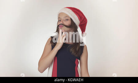 Beautiful happy girl adolescent dans un santa claus hat émotionnellement fools autour et fait une moustache hors de ses cheveux sur fond blanc Banque D'Images