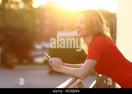 Side view portrait of a happy girl thinking et tenant un téléphone intelligent d'une maison ou d'un balcon de l'hôtel au coucher du soleil Banque D'Images