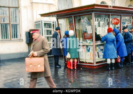 Scène dans une rue de Moscou de gens faisant la queue devant un kiosque à journaux, novembre 1973. Des panneaux en russe annoncent la vente de journaux et de magazines. Au premier plan, un homme en uniforme marche avec une mallette à la main. () Banque D'Images