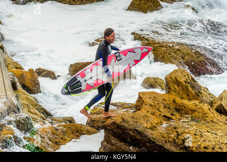 Un surfeur étapes soigneusement sur les rochers et dans des conditions de surf dangereux à Bronte Beach à Sydney, NSW, Australie Banque D'Images