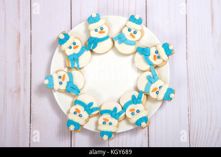 Le SNOWMAN sugar cookies décorés de marshmallow fondant, le glaçage royal bleu foulard et chapeau disposées en cercle sur une assiette de porcelaine de table en bois clair. Banque D'Images