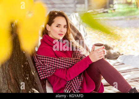 Cute femme s'asseoir sur un banc de parc en automne, holding cup et à la caméra à l'extérieur shot. Banque D'Images