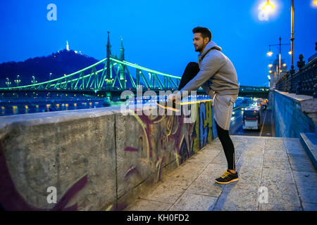 Un jeune sportif séduisant associe sa cirage sur la rue la nuit au bord de la rivière avec un pont derrière lui Banque D'Images