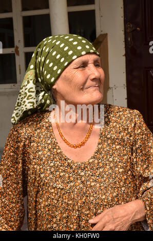 Vieille dame fière, Babushka, avec une écharpe sur la tête, collier et robe avec motif, est souriant / riant. Dans un petit village au Tadjikistan. Banque D'Images