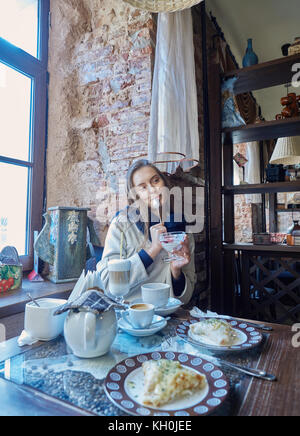 Une charmante jeune fille aux cheveux noirs se repose dans un café confortable. Le café est sur sa table. La jeune fille aime le goût de la crème glacée. Banque D'Images