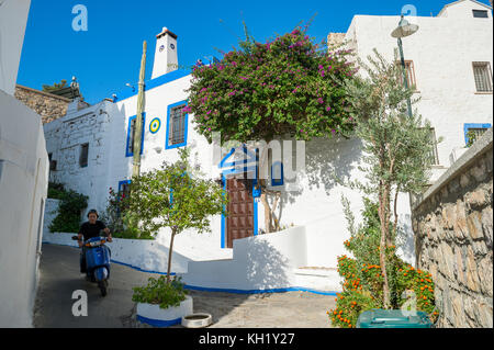 BODRUM, TURQUIE - 6 OCTOBRE 2016 : un homme en scooter traverse une ruelle étroite d'architecture méditerranéenne bleue et blanche traditionnelle avec bougainvil Banque D'Images