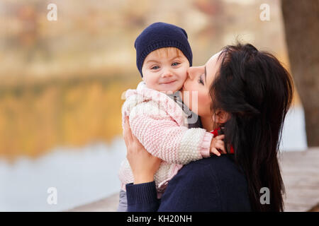 Jeune mère de baisers petite fille dans la forêt d'automne près de la rivière. maman holding baby girl à mains. famille heureuse, l'amour parental, saison automne concep Banque D'Images