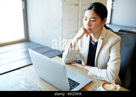 Jeune femme sérieuse assis par table à café en face de l'ordinateur portable et la lecture de données Banque D'Images