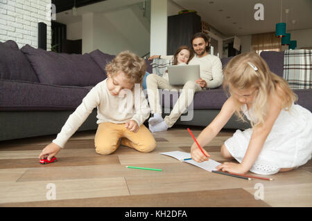 Les frères et sœurs des enfants jouant sur le sol tandis que les parents à l'aide d'ordinateur portable à la maison, preschool boy holding toy voiture, petite fille dessin avec des crayons en album, f Banque D'Images