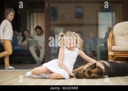 Petite fille jouant avec un chien à l'extérieur, caressant berger allemand allongé sur porche, parents avec petit garçon passer du temps à l'intérieur de la maison à fond, les loisirs Banque D'Images